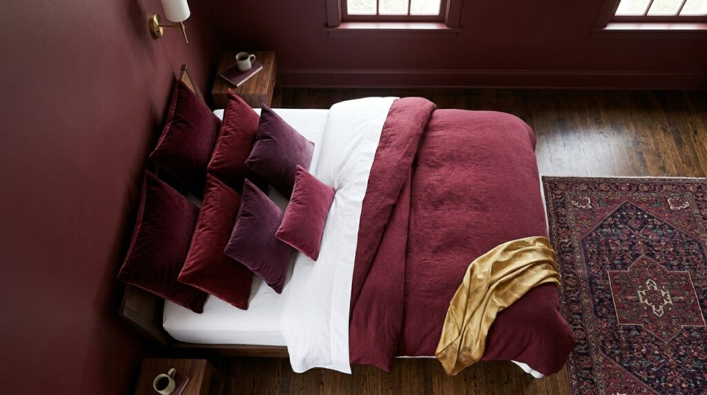 Top-down view of a bed with burgundy velvet pillows and white linens on a dark wood floor with a Persian rug.