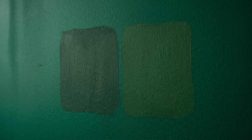 Two dark green paint swatches applied to a textured emerald wall for a bedroom renovation project.