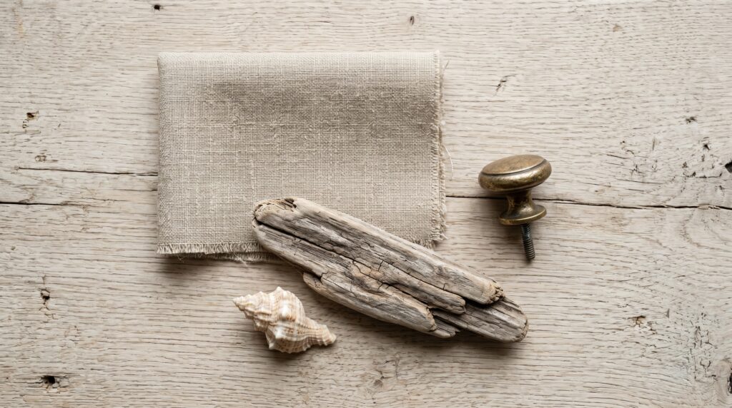 Coastal decor flat lay with driftwood, a seashell, linen fabric, and a brass knob on a rustic light wood background.