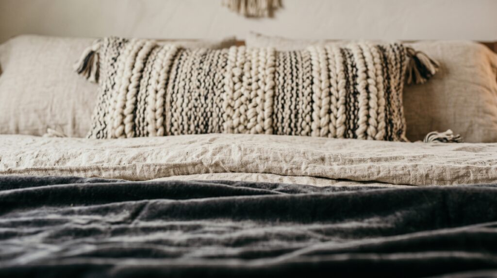 Close-up of neutral bohemian bedding featuring a textured chunky knit lumbar pillow and wrinkled linen sheets on a bed.